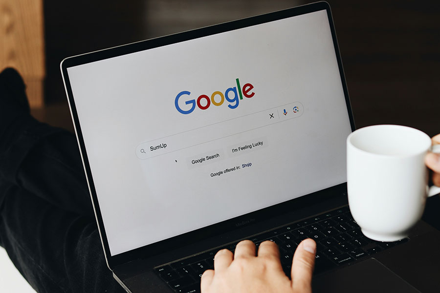 Google search engine open on a laptop, hands over the laptop typing and holding a mug.