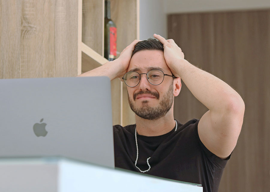 A man staring at a laptop with his hands on his head, looking like he’s about to cry.